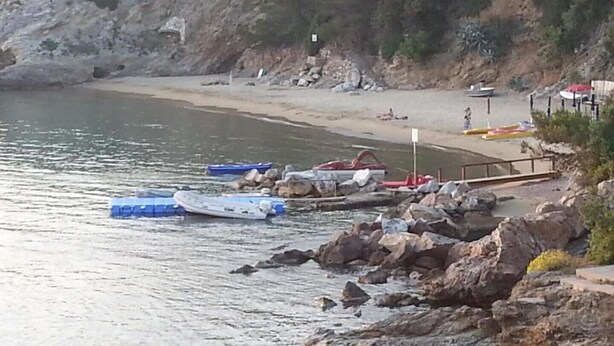 Une côte rocheuse bordée de bateaux amarrés près de l’eau, une plage de sable et une falaise couverte de végétation.