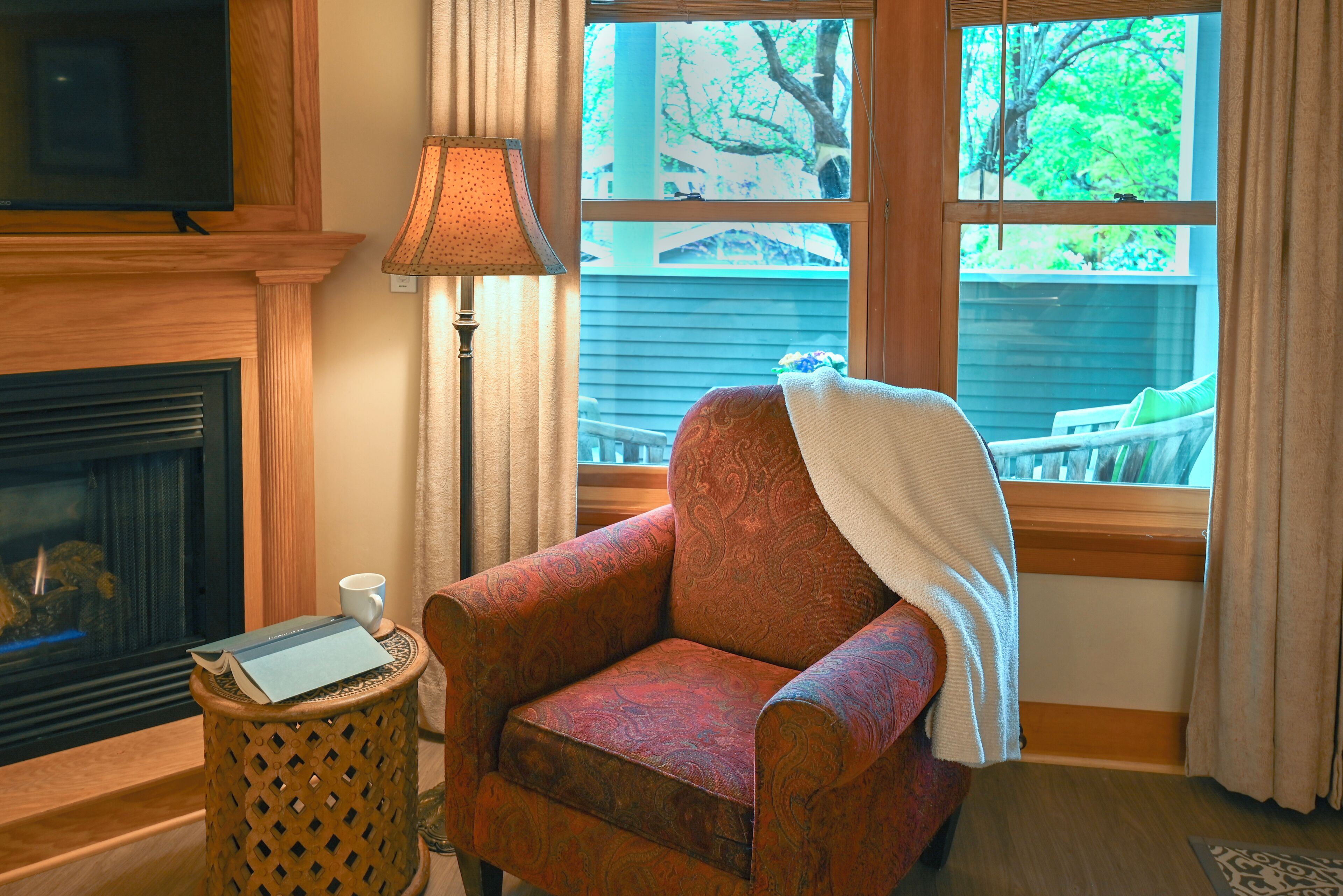 A cozy living room with a fireplace, a comfortable armchair, a small side table with a book, and a view of the outdoors through large windows.