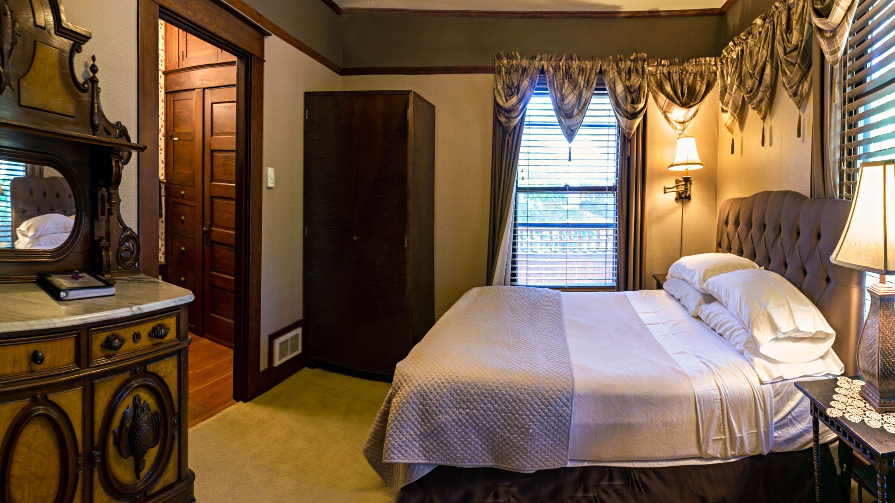 A bedroom with a large bed, a dresser, a mirror, a wardrobe, and a window with curtains.