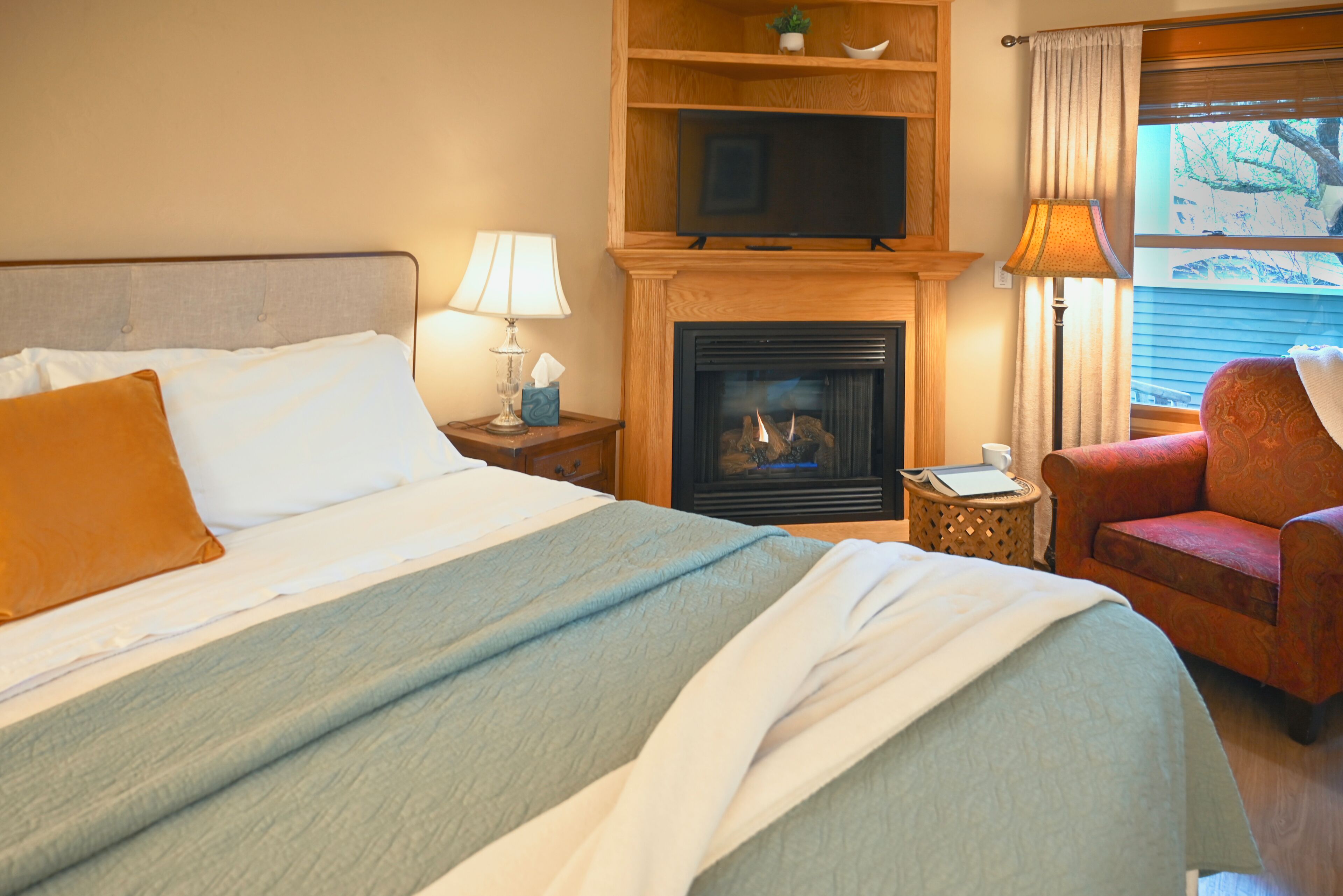 A hotel room with a bed, a television, a fireplace, a lamp, a chair, and a window with a view.