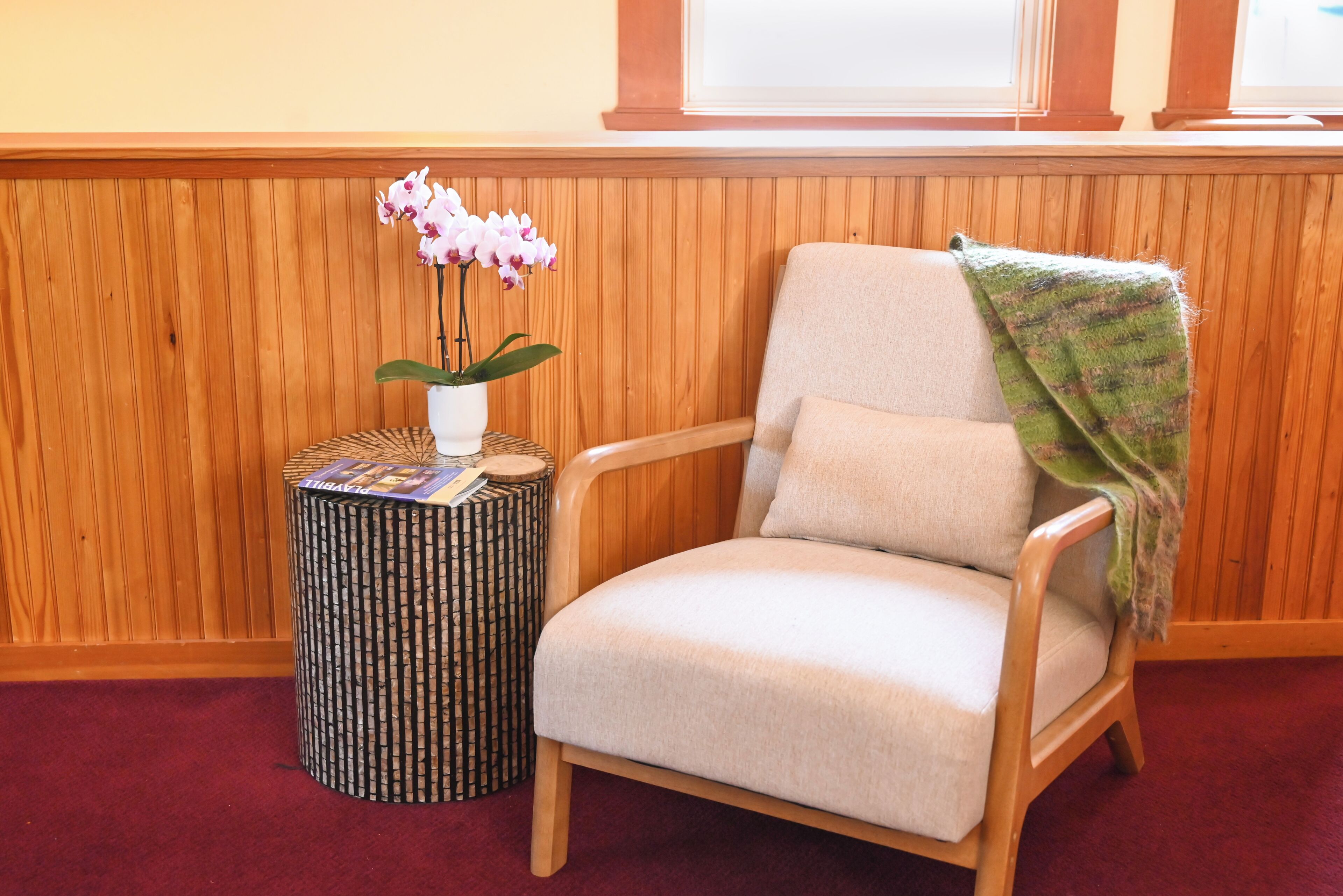 Une pièce aux murs lambrés, avec un fauteuil beige, une petite table d’appoint ornée d’une plante et un tapis à motifs.