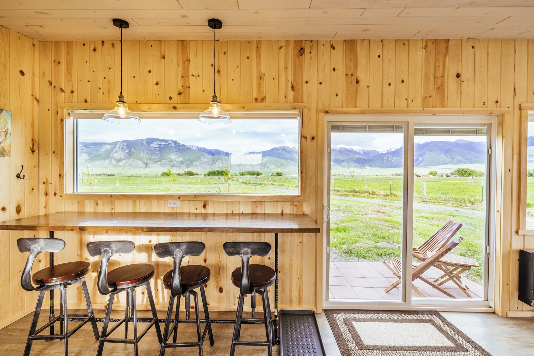 A wooden interior with a bar area, four stools, and a large window offering a mountain view.