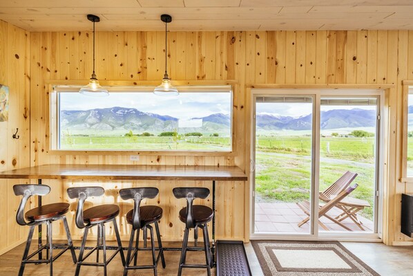 Interior de madera con una zona de bar, cuatro taburetes y un amplio ventanal con vistas a la montaña.