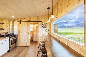 A wooden interior with a kitchen, dining area, and large windows offering a view of a grassy field and mountains.