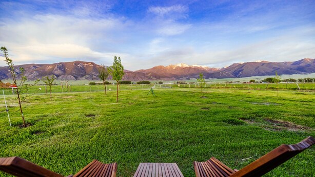 A grassy field with two wooden chairs and a table, surrounded by mountains and a clear sky.