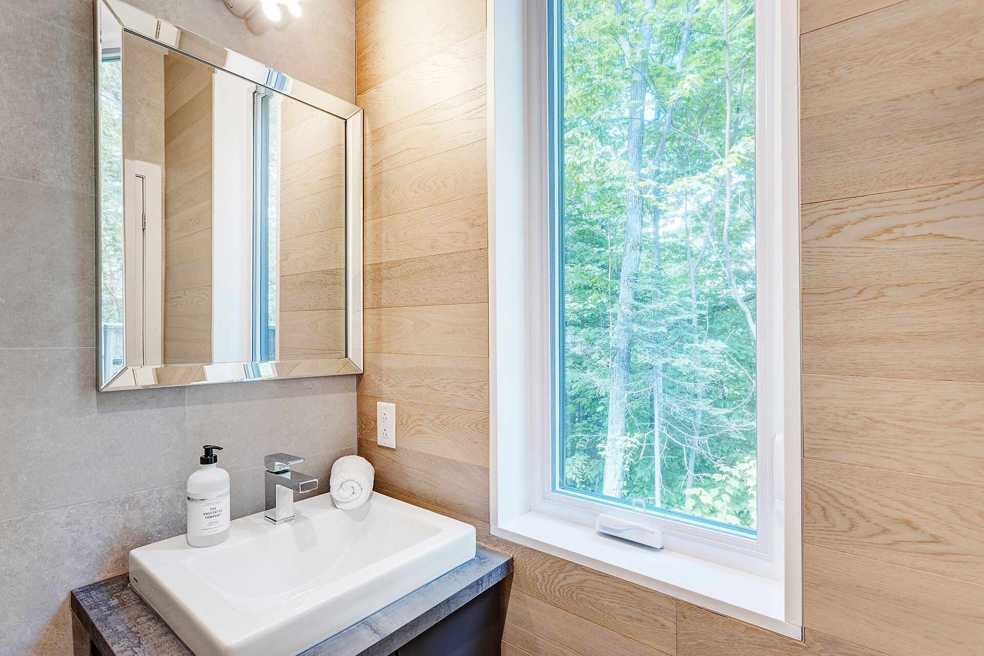A bathroom with a white sink, a mirror, and a window with a view of trees.