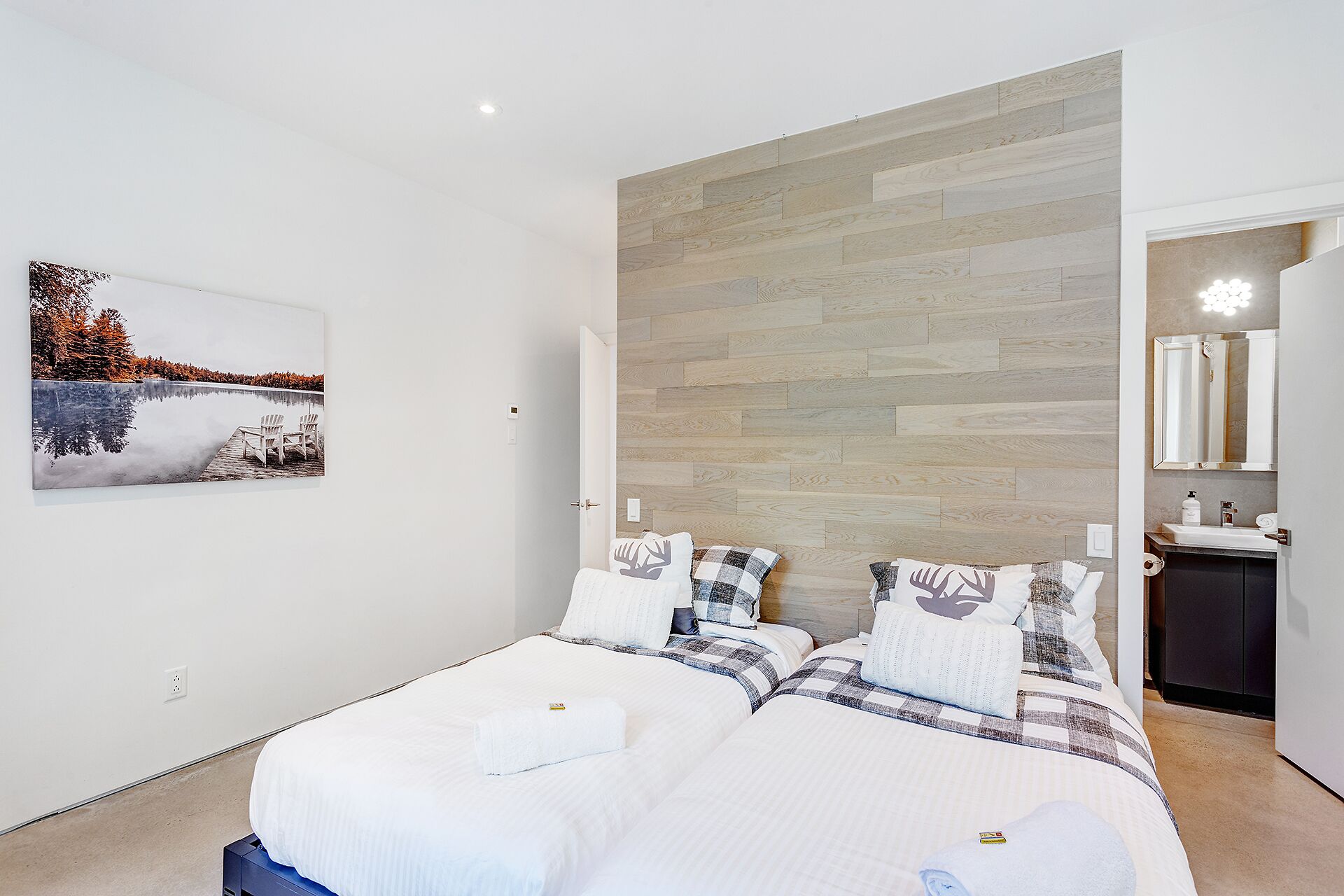 Un dormitorio con dos camas, una pared de acento de madera, un baño con espejo y lavamanos, y un cuadro que representa una escena junto al lago.