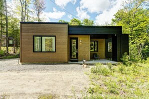A modern wooden cabin with a large window, a door, and a porch area. - Tremblant Oasis: Luxury, Nature, Hot Tub, Views! (La Conception)