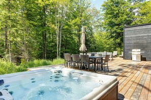 A hot tub with a wooden deck, outdoor furniture, and a wooden building in a forested area. - Tremblant Oasis: Luxury, Nature, Hot Tub, Views! (La Conception)