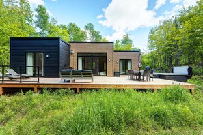 A modern house with a deck, outdoor seating, and a large window overlooking a forest. - Tremblant Oasis: Luxury, Nature, Hot Tub, Views! (La Conception)