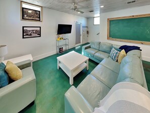 A room with a green floor, a white table, a light blue sofa, and a television on a stand. - 7 Heaven by Silver Sands Vacation Rentals (Port Aransas)