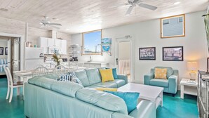 A modern living room with a teal sofa, a coffee table, and a dining area with white furniture. - Above & Beyond by Silver Sands Vacation Rentals (Port Aransas)