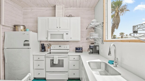 A compact kitchen with white appliances, a sink, and a window with a view of palm trees. - Above & Beyond by Silver Sands Vacation Rentals (Port Aransas)