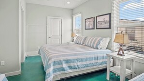 A bedroom with a bed, a ceiling fan, a window with blinds, and a nightstand with a lamp. - Above & Beyond by Silver Sands Vacation Rentals (Port Aransas)