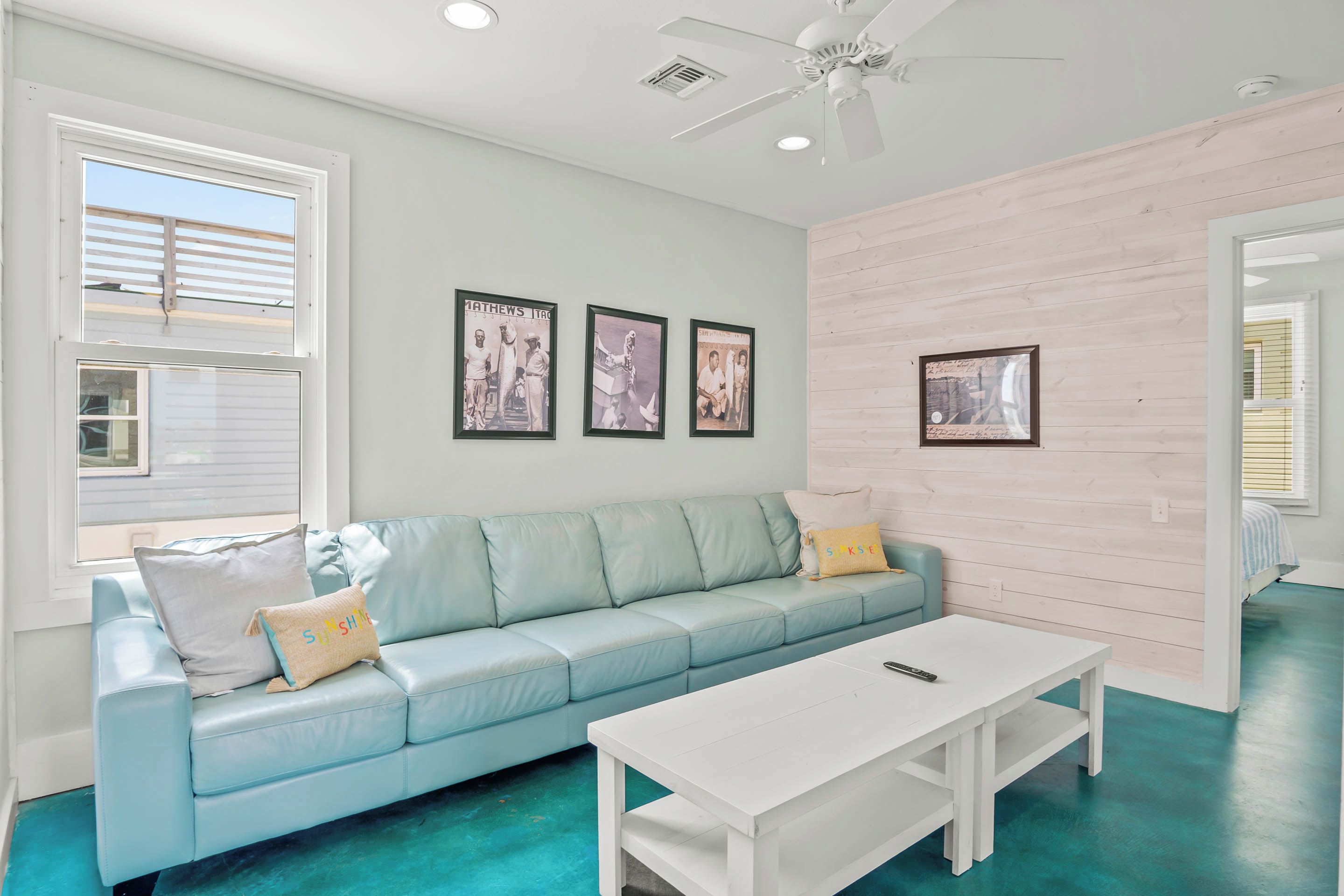 A room with a light blue leather sofa, a white coffee table, and a ceiling fan. There are three framed pictures on the wall and a window with blinds.