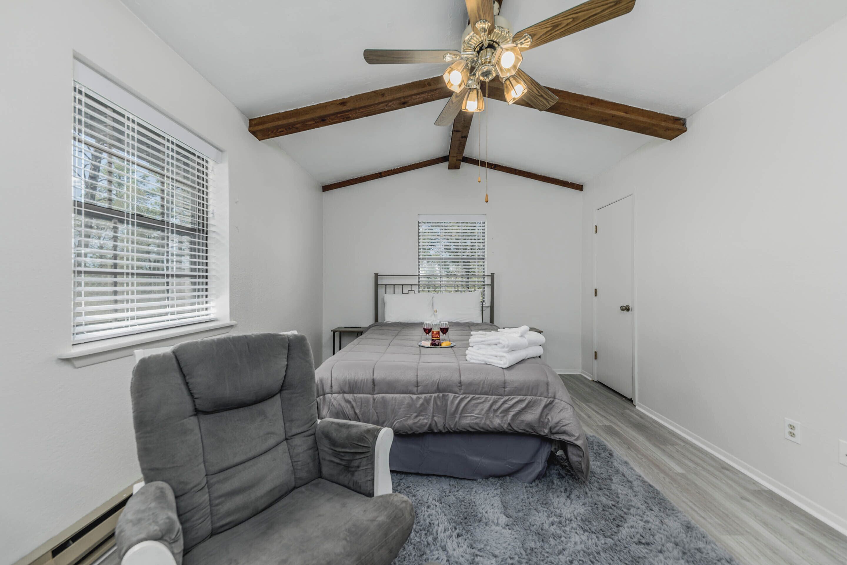 Une chambre à coucher avec un lit, un fauteuil gris, un ventilateur de plafond et une fenêtre avec des stores.