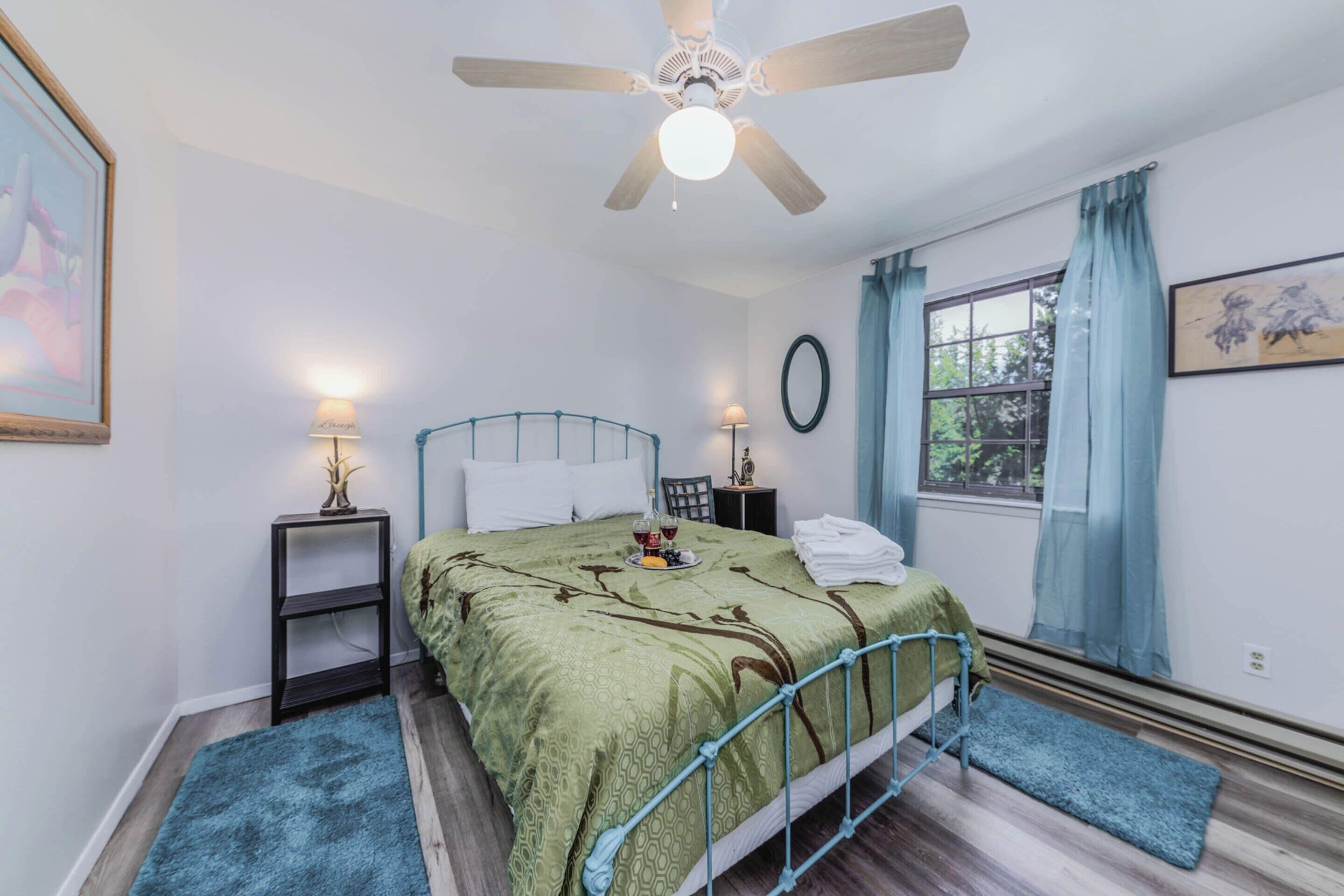 A bedroom with a bed, two bedside tables, a ceiling fan, a window with curtains, and a framed picture on the wall.