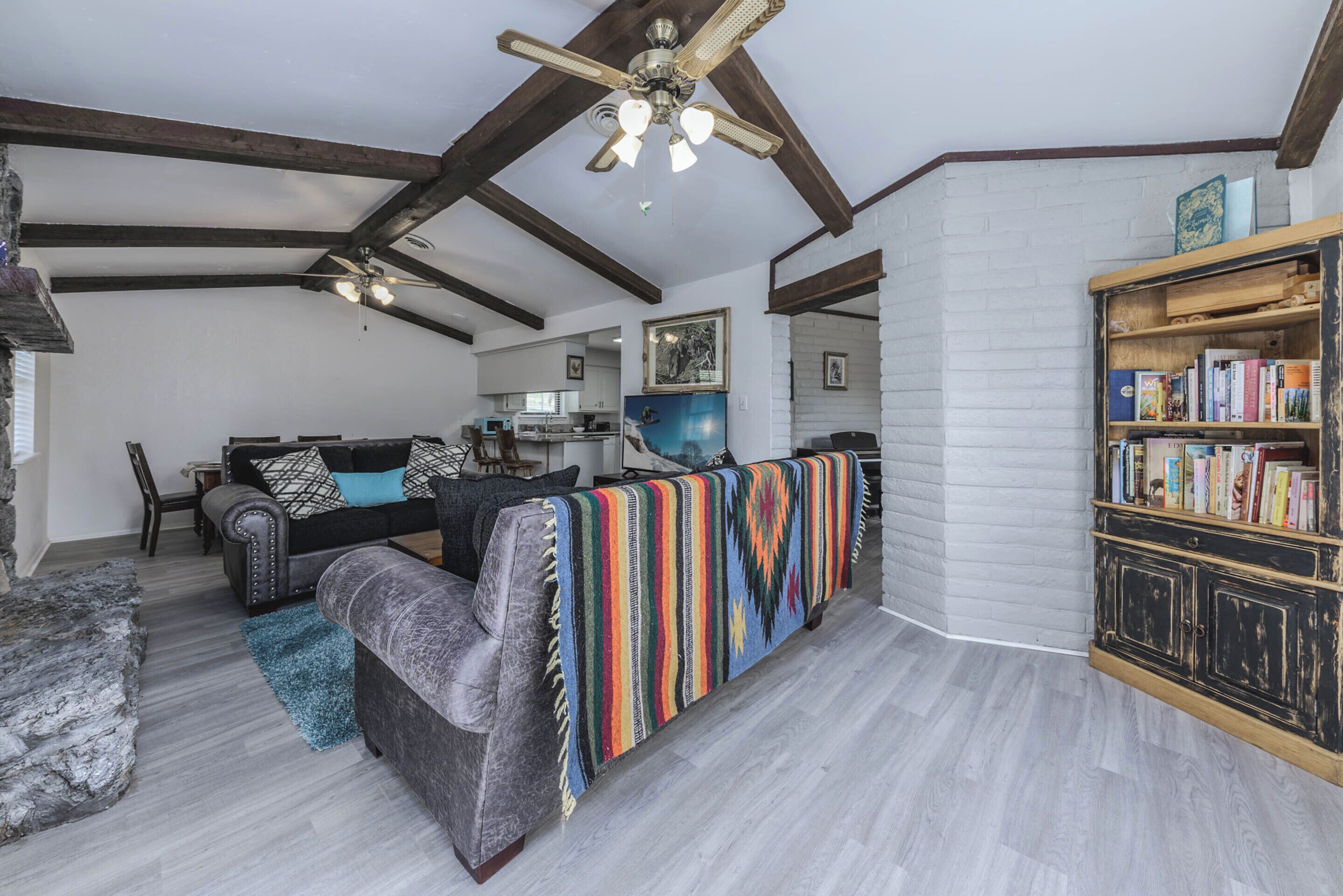 A spacious living room with a high ceiling, exposed wooden beams, a fireplace, a bookshelf, and a cozy seating area with a colorful blanket.