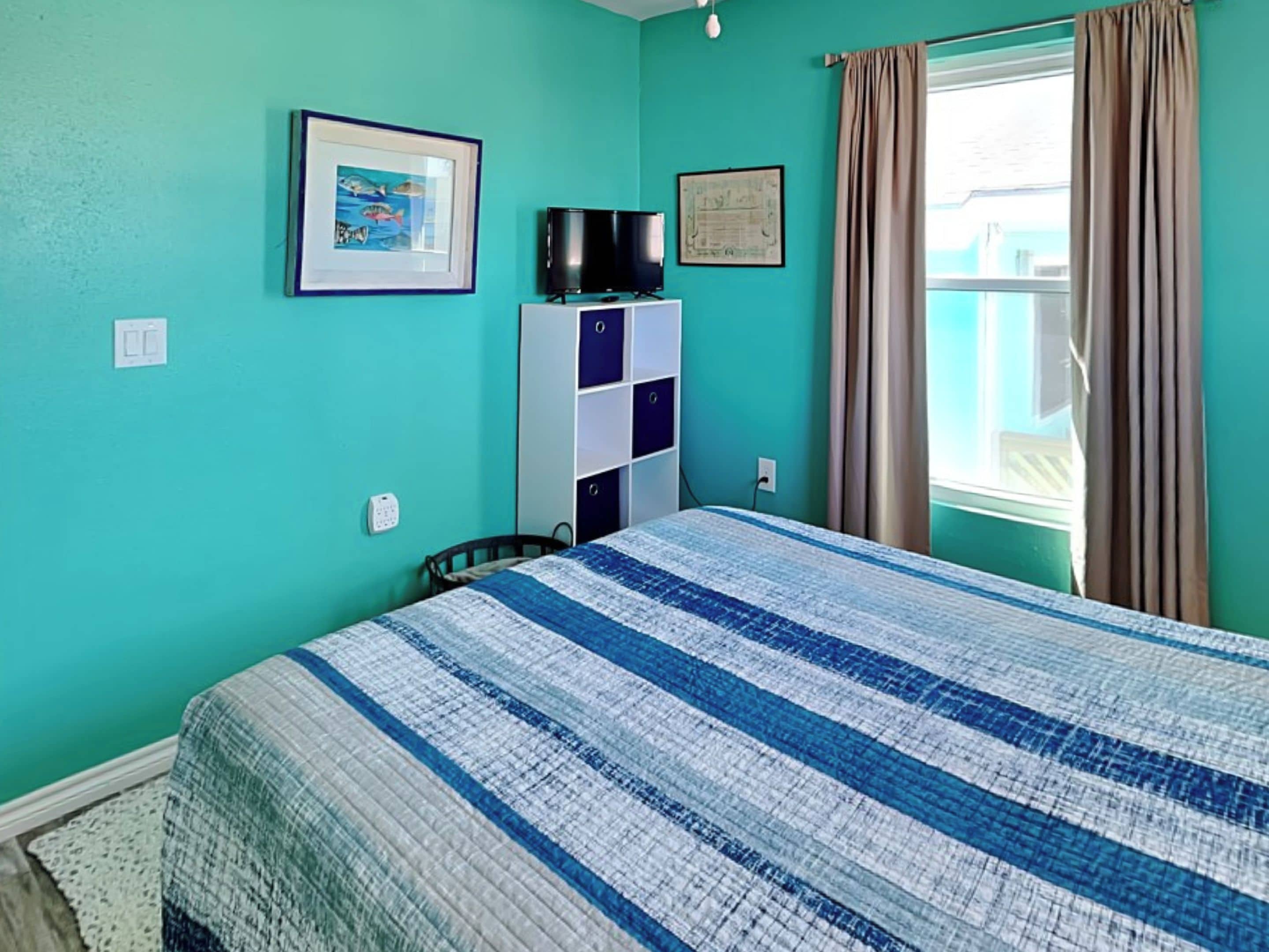 Une chambre avec des murs de couleur turquoise, un lit recouvert d’une couette à rayures bleues et blanches, un téléviseur posé sur un meuble blanc, un tableau encadré accroché au mur et une fenêtre avec des rideaux.