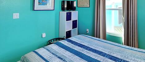 Une chambre avec des murs de couleur turquoise, un lit recouvert d’une couette à rayures bleues et blanches, un téléviseur posé sur un meuble blanc, un tableau encadré accroché au mur et une fenêtre avec des rideaux.