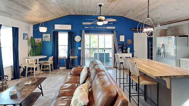 A living room with a brown leather sofa, a wooden coffee table, and a kitchen area with a wooden countertop and bar stools. - Sand Piper by Miss Kitty’s (Rockport)