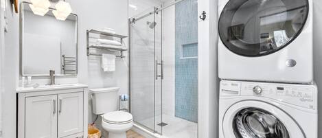 Een moderne badkamer met een douchecabine van glas, een ingebouwde wasmachine en een witte wastafel met spiegel.