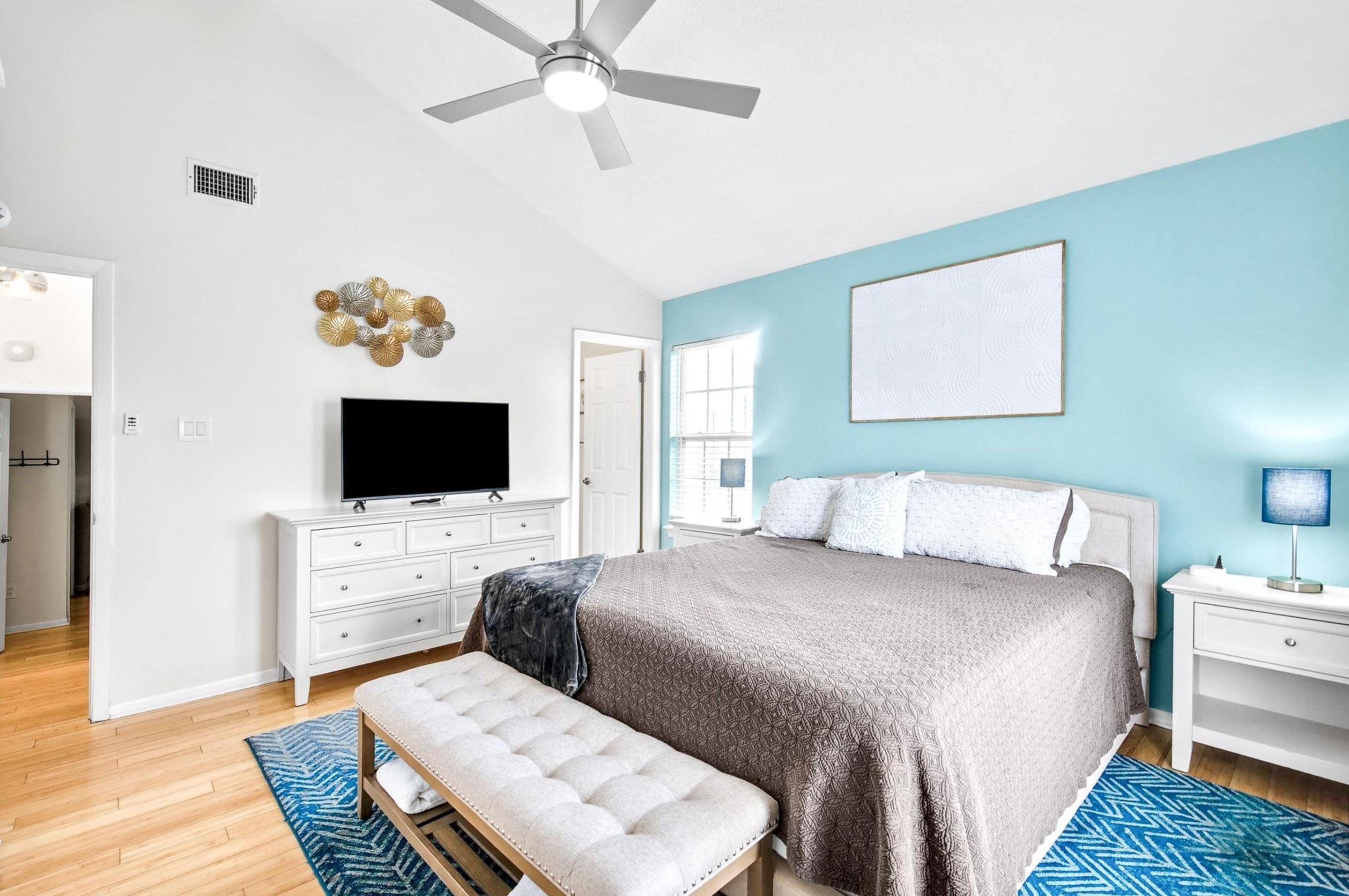 A bedroom with a bed, a TV, a ceiling fan, a bench, and a blue rug.