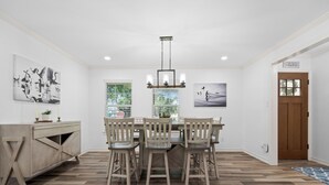 A dining area with a wooden table and chairs, a light fixture above, and artwork on the walls. - Reel Blessing by Miss Kitty’s (Rockport)