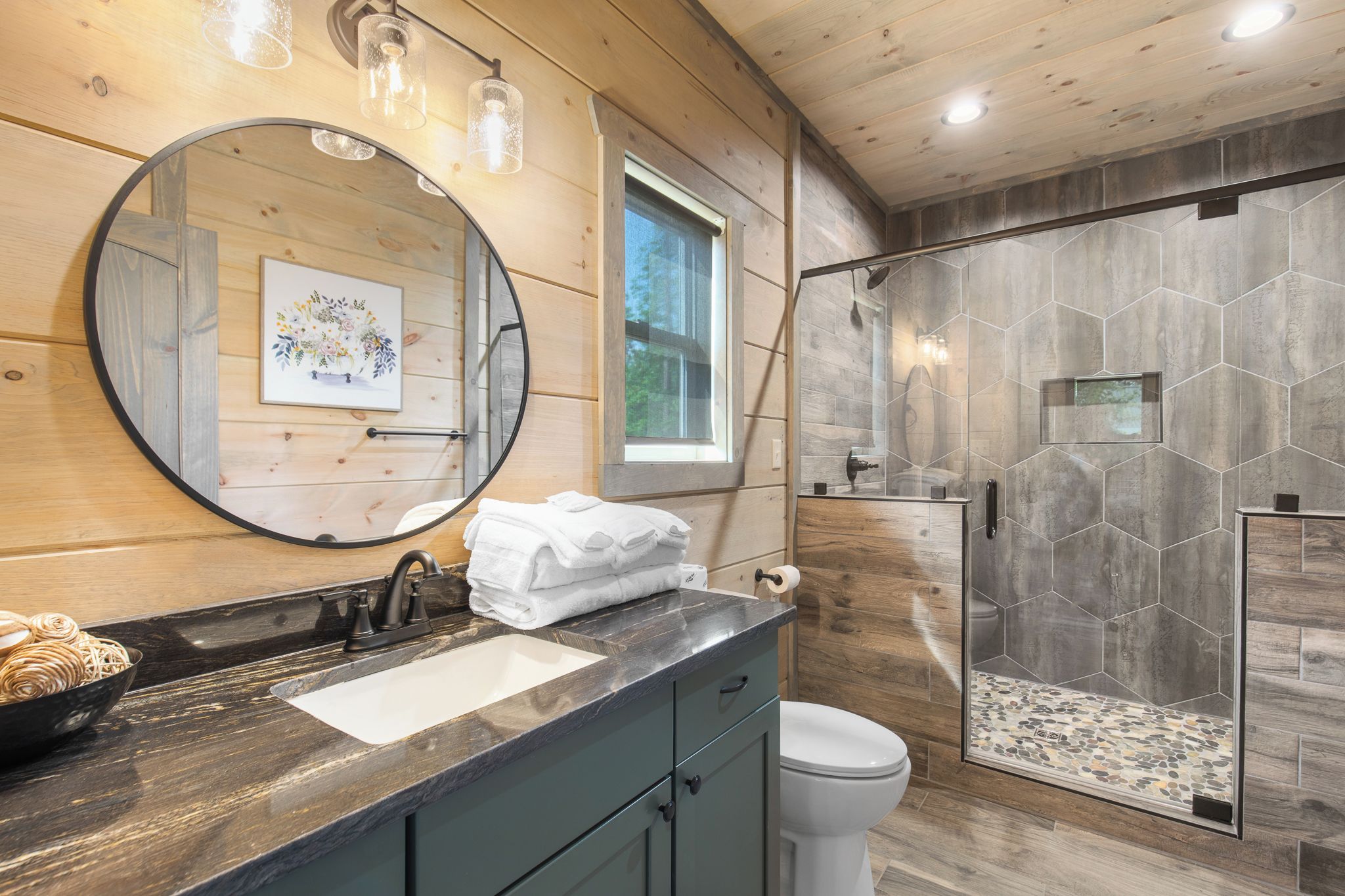 A bathroom with a wooden interior, a round mirror, a sink with a marble countertop, and a shower with hexagonal tiles.