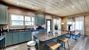 A modern kitchen with wooden ceiling beams, a large island with stools, and a view of the outdoors through a window.