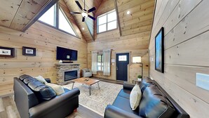 A log cabin interior with two black leather sofas, a stone fireplace, a ceiling fan, and a television mounted on the wall.