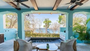 A sunlit patio with wicker furniture, a small table, and a view of a water feature and trees. - Casa de los Sueños- 5 BR Home at Las Catalinas (Catalinas)