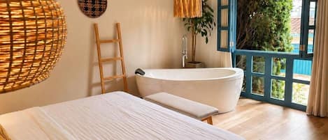 A bedroom with a large bed, a bathtub, a wooden ladder, and a view through an open door.