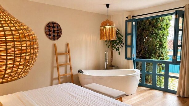 A bedroom with a large bed, a bathtub, a wooden ladder, and a view through an open door.