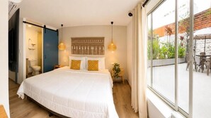 A bedroom with a large bed, a bedside table, a hanging light fixture, and a view of the outdoors through a window.