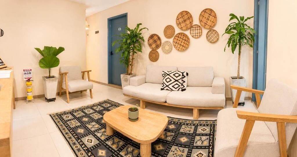 A living room with a sofa, two armchairs, a coffee table, and a patterned rug. There are two potted plants and a wall-mounted decorative item.