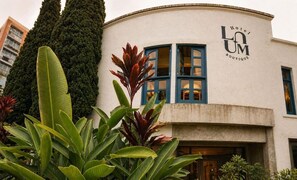 Um edifício com fachada curva, um letreiro com a inscrição 'Hotel LUM Boutique' e uma grande janela com persianas azuis.