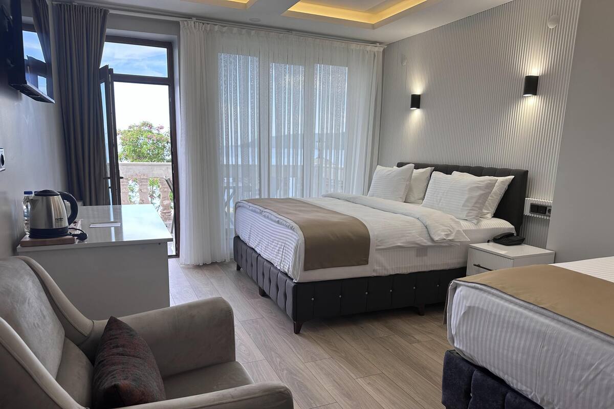 A modern hotel room with a bed, a sofa, a small table with a kettle, and a view of the outdoors through large windows.