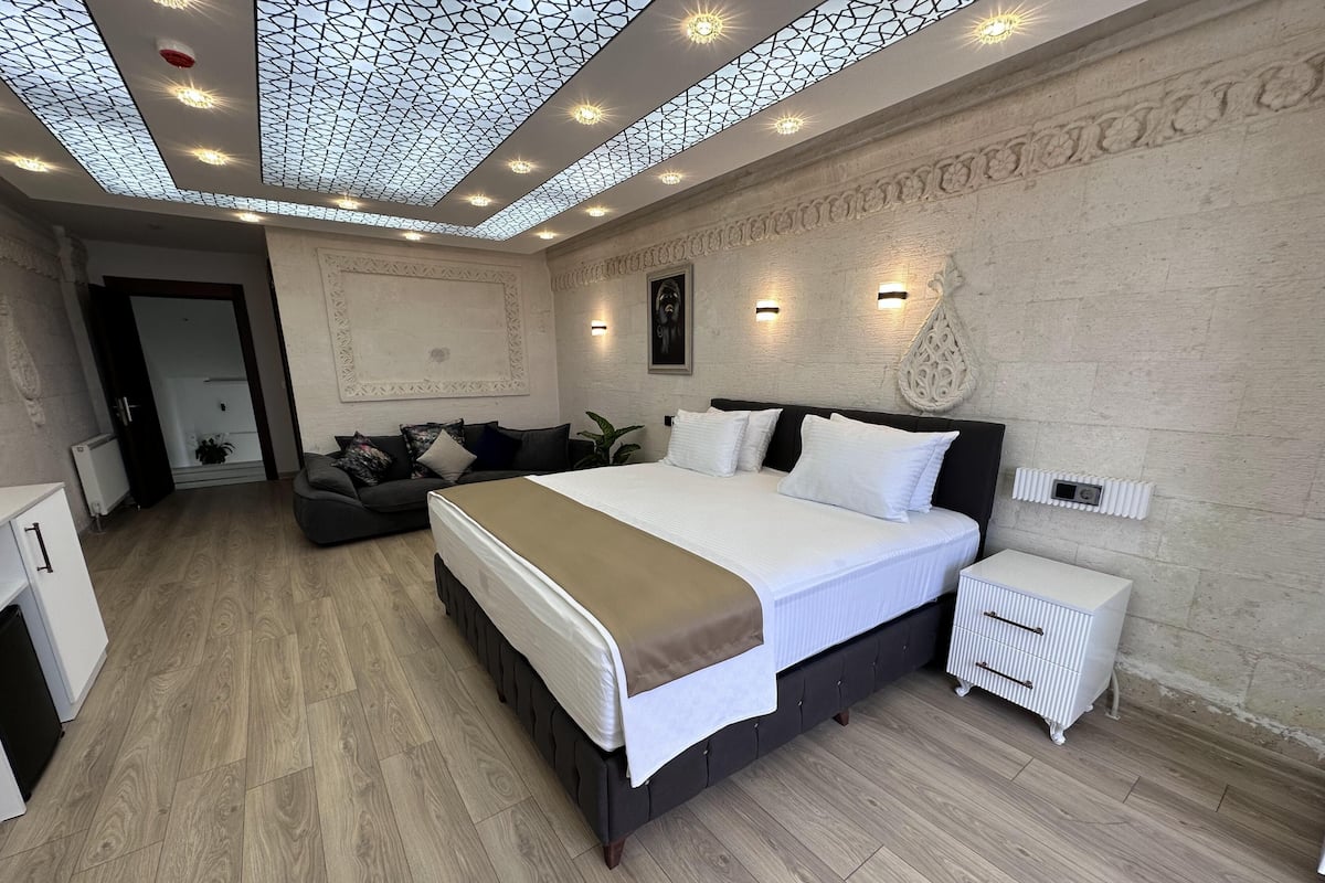 A modern bedroom with a large bed, a dark-colored sofa, a small white bedside table, and a decorative ceiling.