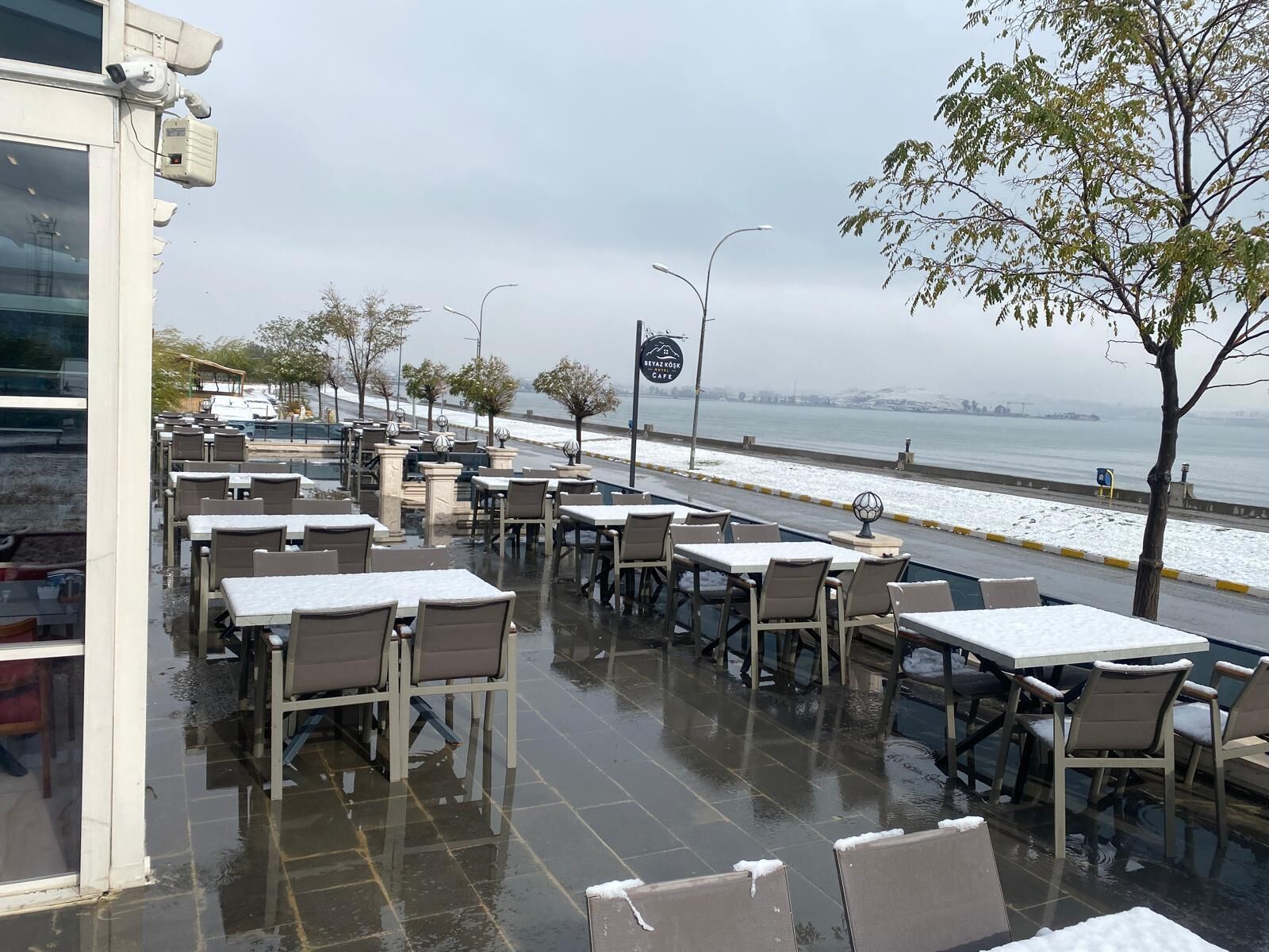 An outdoor cafe with tables and chairs, a view of the sea, and a cafe sign.