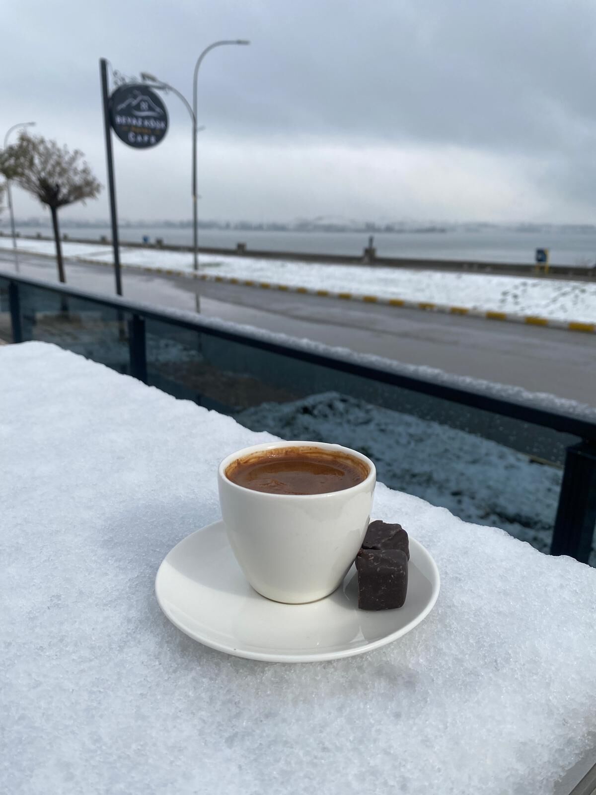 A cup of coffee and a chocolate piece on a snow-covered surface with a view of a road and a sign in the background.
