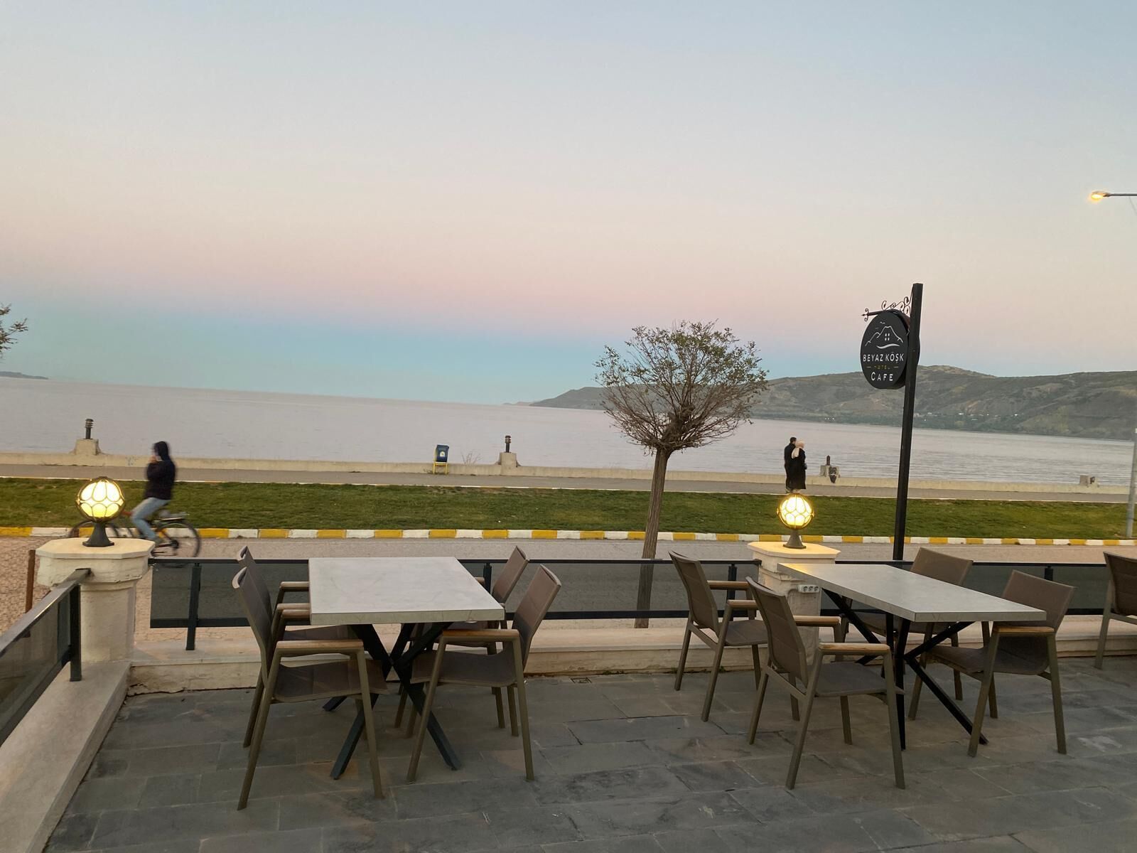 An outdoor seating area with tables and chairs, overlooking a body of water and a hilly landscape.