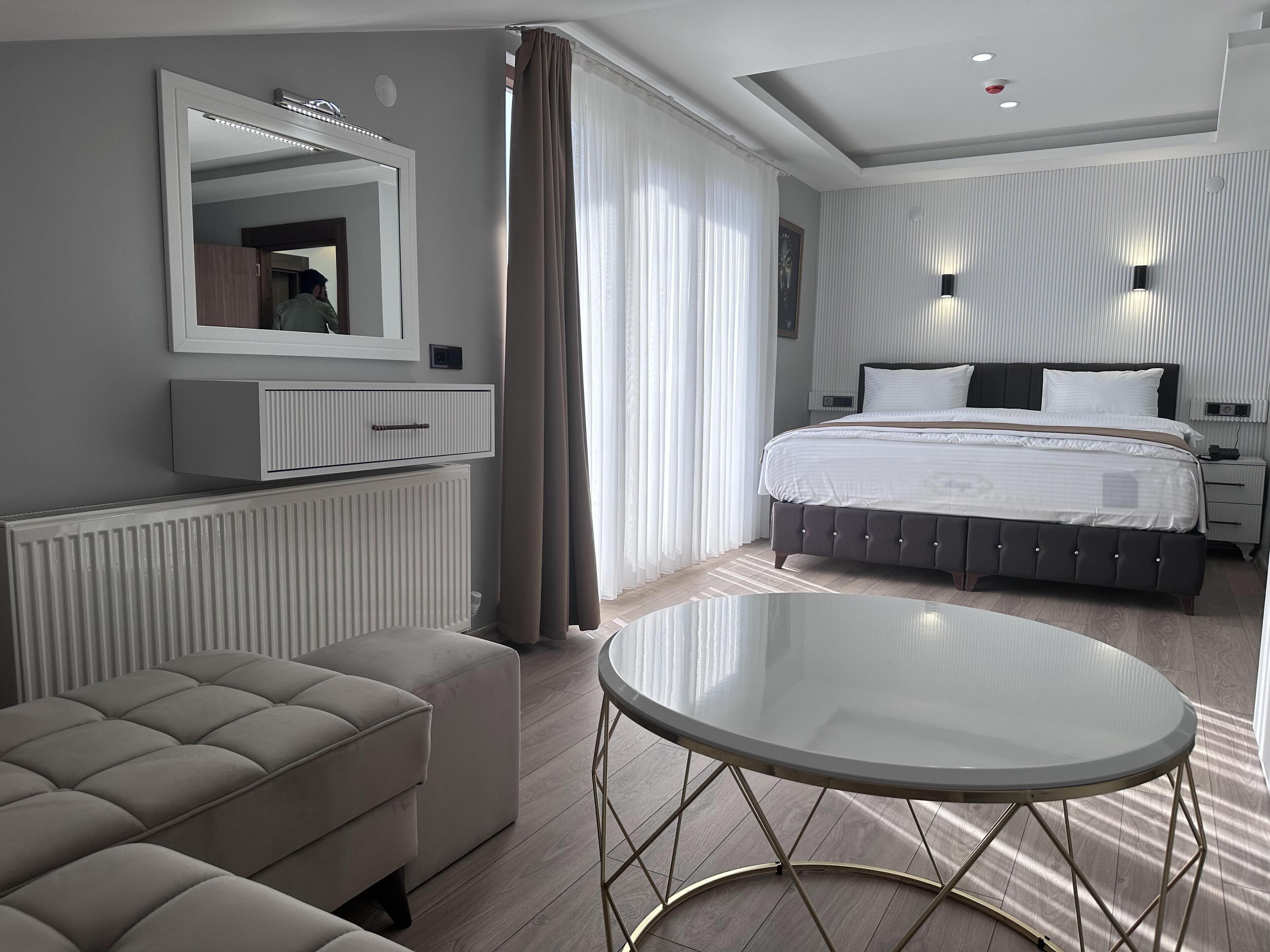 A modern hotel room with a large bed, a round glass table, a sofa, and a mirrored wardrobe.