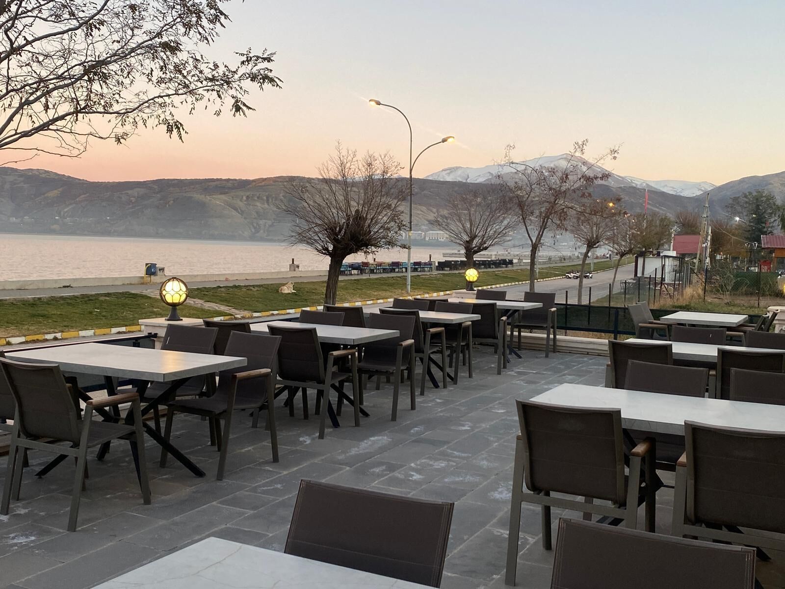 An outdoor dining area with tables and chairs, a lamp post, and a view of a body of water and mountains in the background.