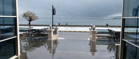 An outdoor seating area with tables and chairs, a view of the sea, and a snow-covered ground.