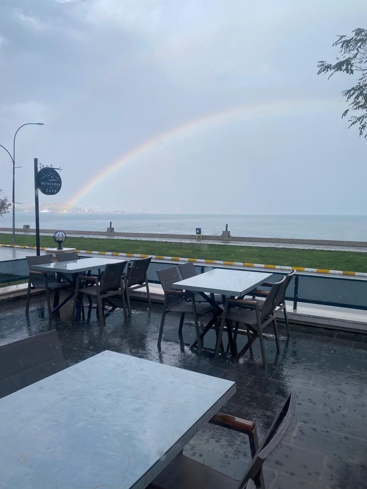 An outdoor cafe with tables and chairs, a sign for BeyazKosk Cafe, and a view of the sea with a rainbow.