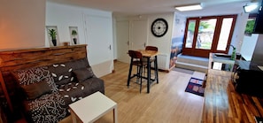 A living room with a sofa, a small table, a wooden dining table with chairs, a clock on the wall, and a kitchen area with a sink and countertop.