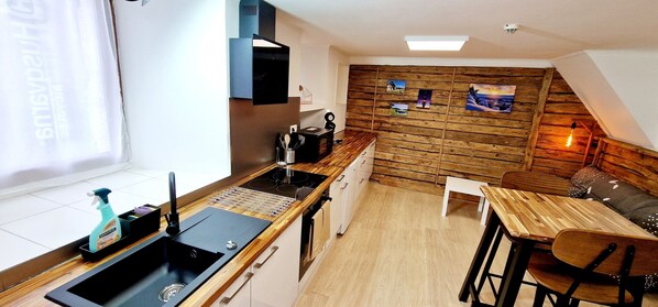 A compact kitchen with a modern stove, a sink, and a wooden wall with pictures. There's a small dining area with a table and chairs.
