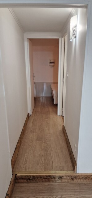 A narrow hallway with wooden flooring leading to a room with a bed and a small table.