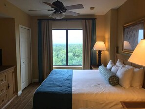 Habitación de hotel con cama, ventilador de techo, ventana con cortinas, lámpara y un cuadro en la pared.